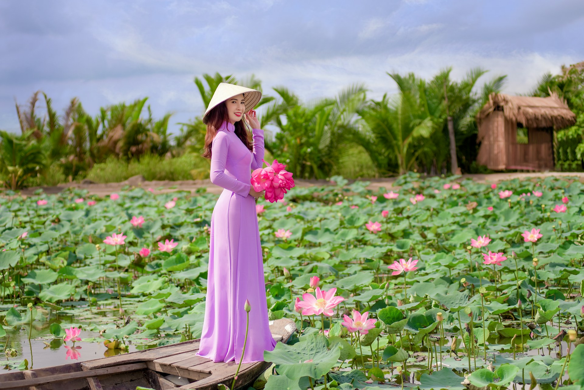 Vietnamese woman in a purple Áo dài and conical hat, standing on a boat surrounded by pink lotus flowers.
