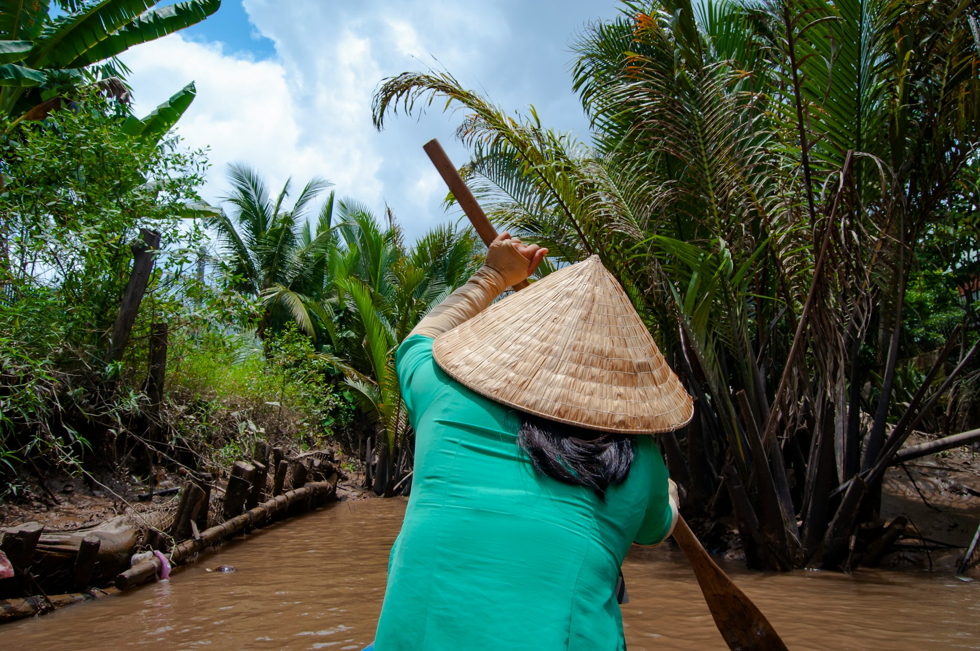 A woman wearing a traditional woven nón lá conical hat rows a boat through a lush tropical Vietnamese waterway.