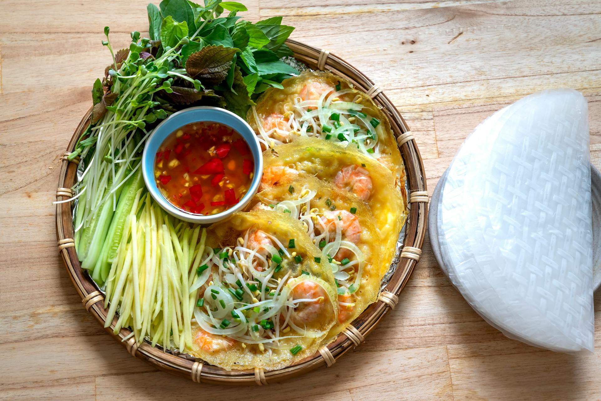 Crispy golden Vietnamese Bánh xèo crepes filled with shrimp, served on bamboo with fresh herbs, greens, and dipping sauce.
