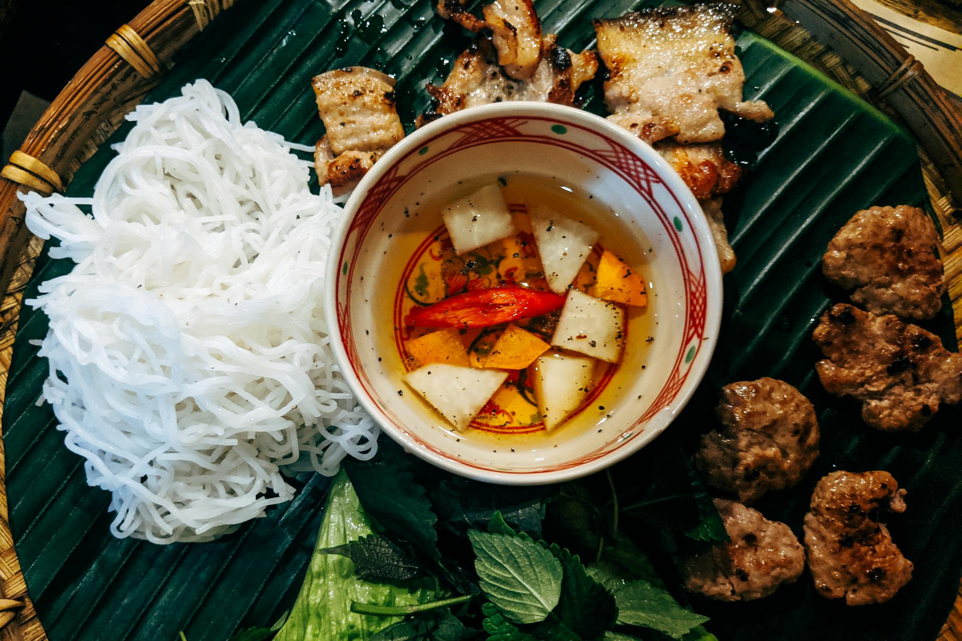 Traditional Vietnamese Bún chả featuring charred pork, white rice vermicelli, fresh herbs, and dipping sauce on a banana leaf.