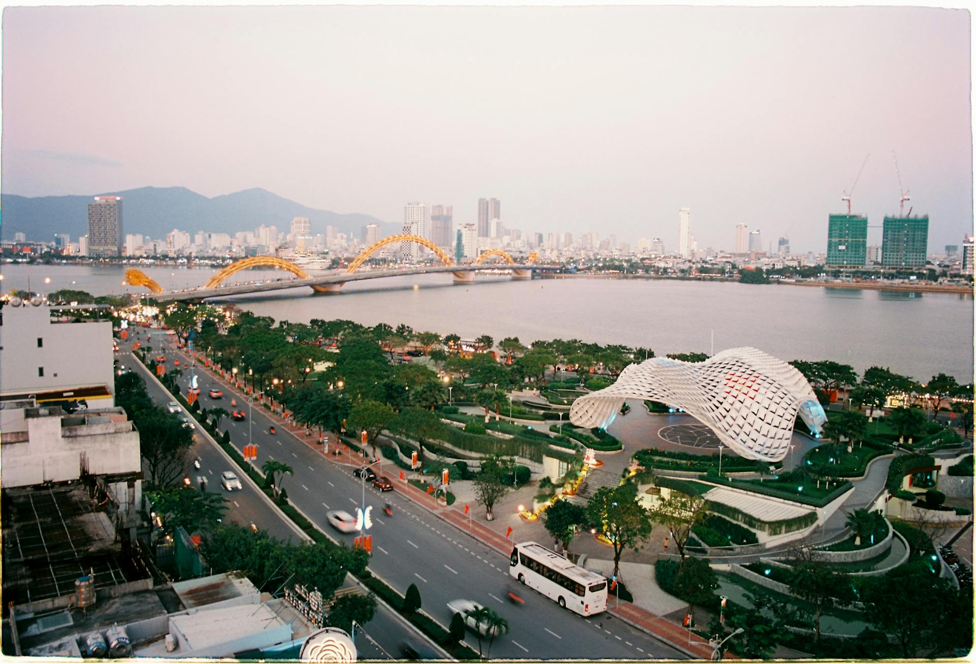 Aerial view of Da Nang's golden Dragon Bridge and Han River, featuring modern city skyscrapers and hazy blue mountains.