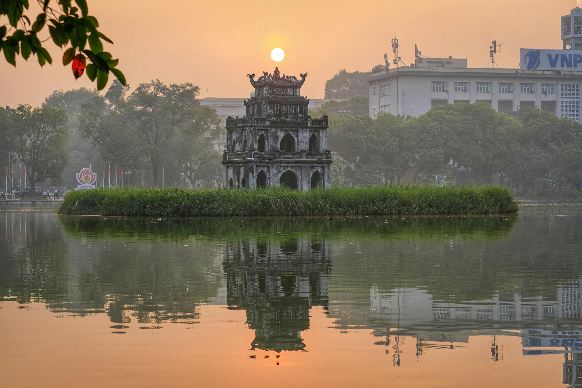Iconic Turtle Tower on Hanoi's Hoan Kiem Lake at sunset, reflecting ancient stone architecture in calm, orange-hued water.