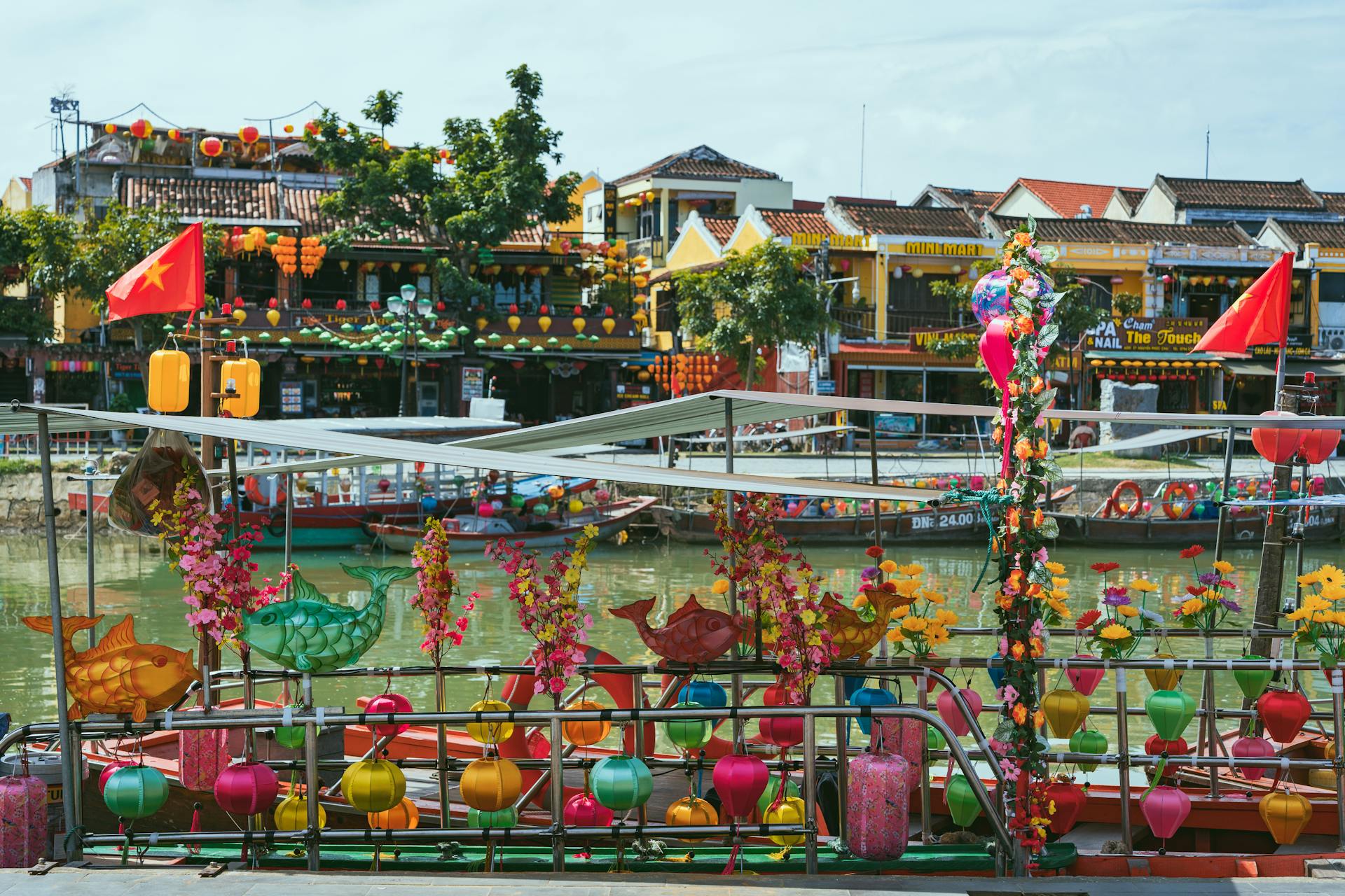 Colorful silk lanterns and fish ornaments on a riverboat, overlooking the iconic yellow heritage buildings of Hội An.