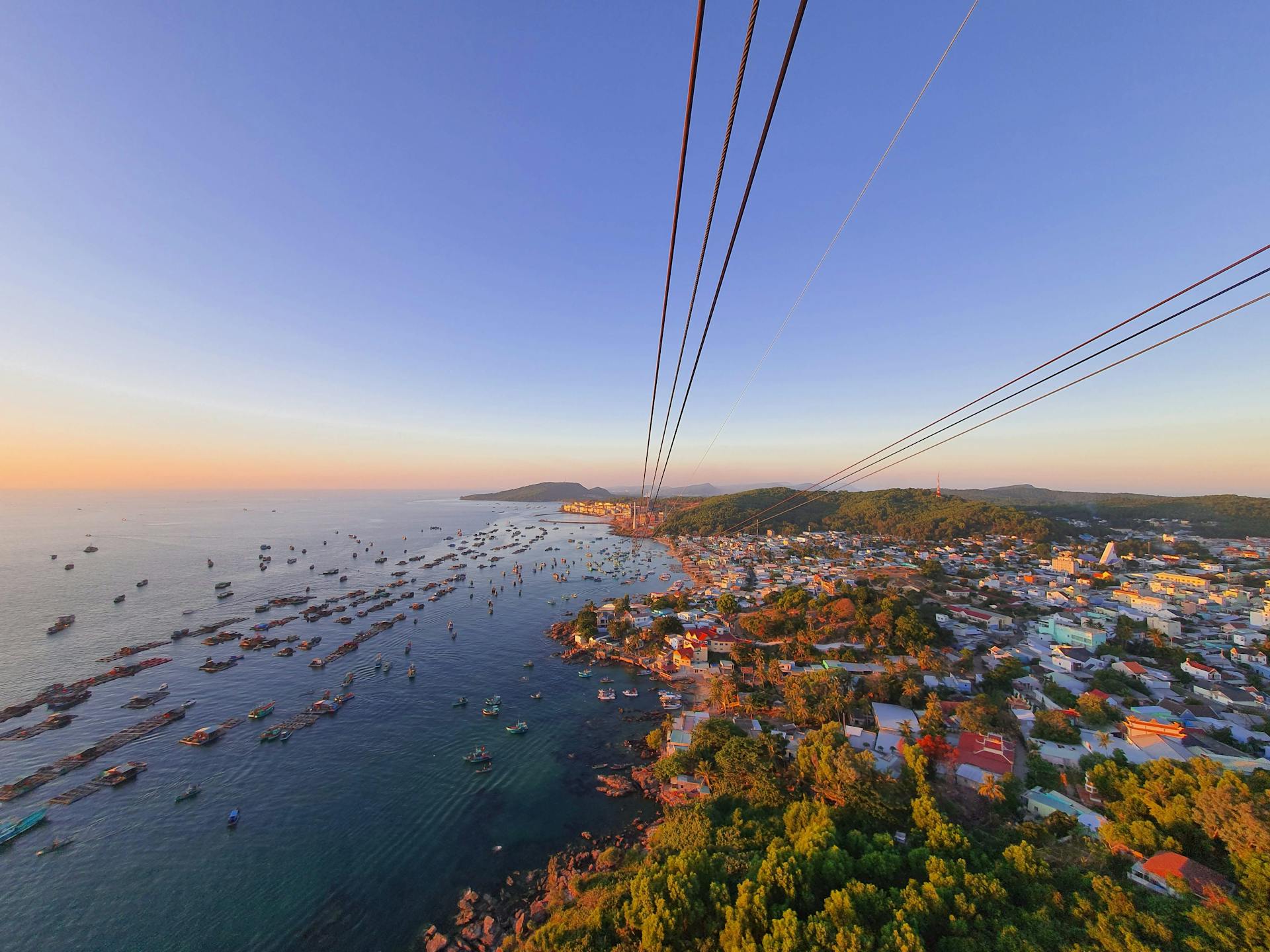 Phú Quốc's coastline with turquoise water, colorful fishing boats, and red-roofed houses seen from the high-altitude Hon Thom cable car.
