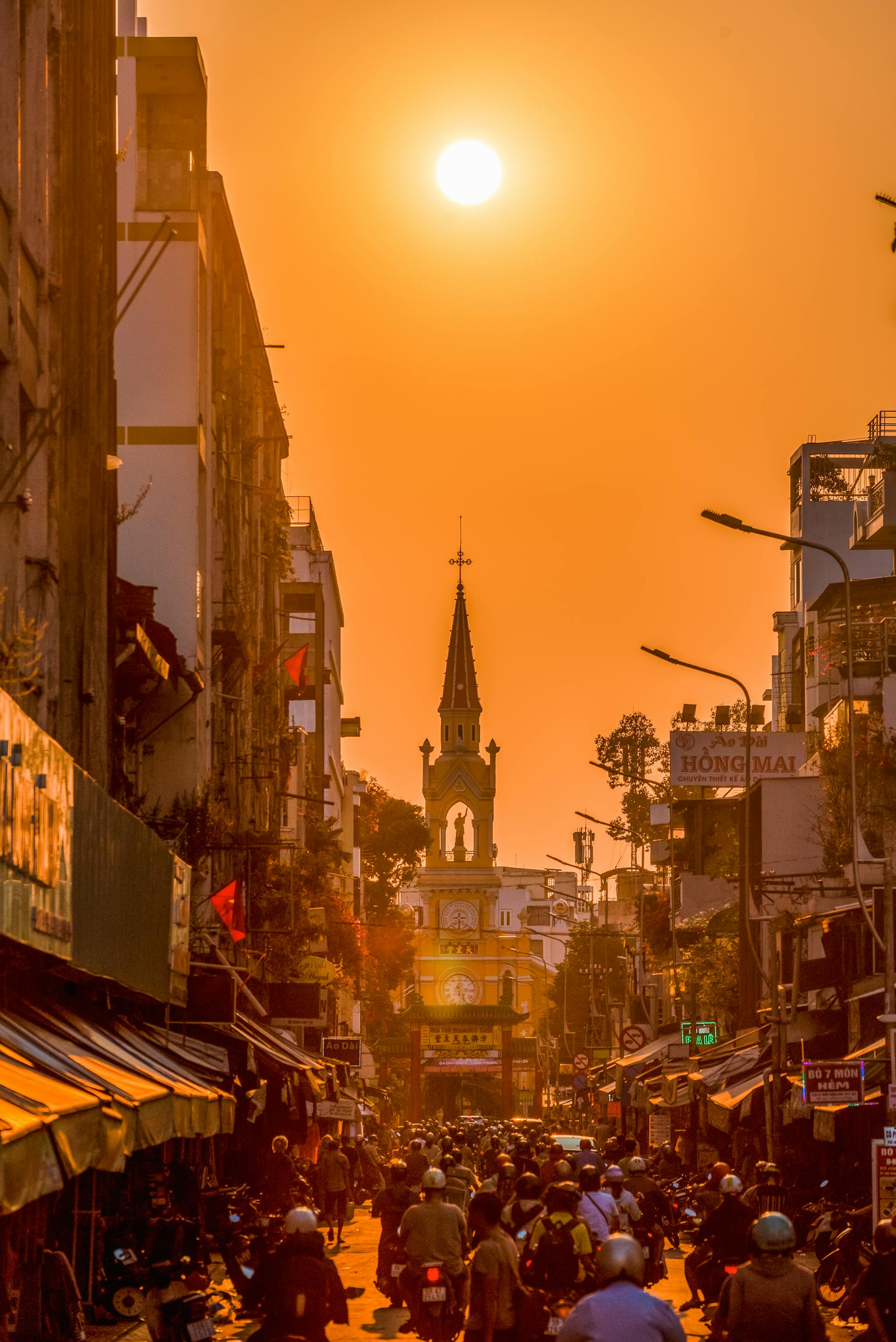 Bustling Sài Gòn street crowded with motorbikes at golden hour, featuring the historic Cha Tam Church spire under a sunset.